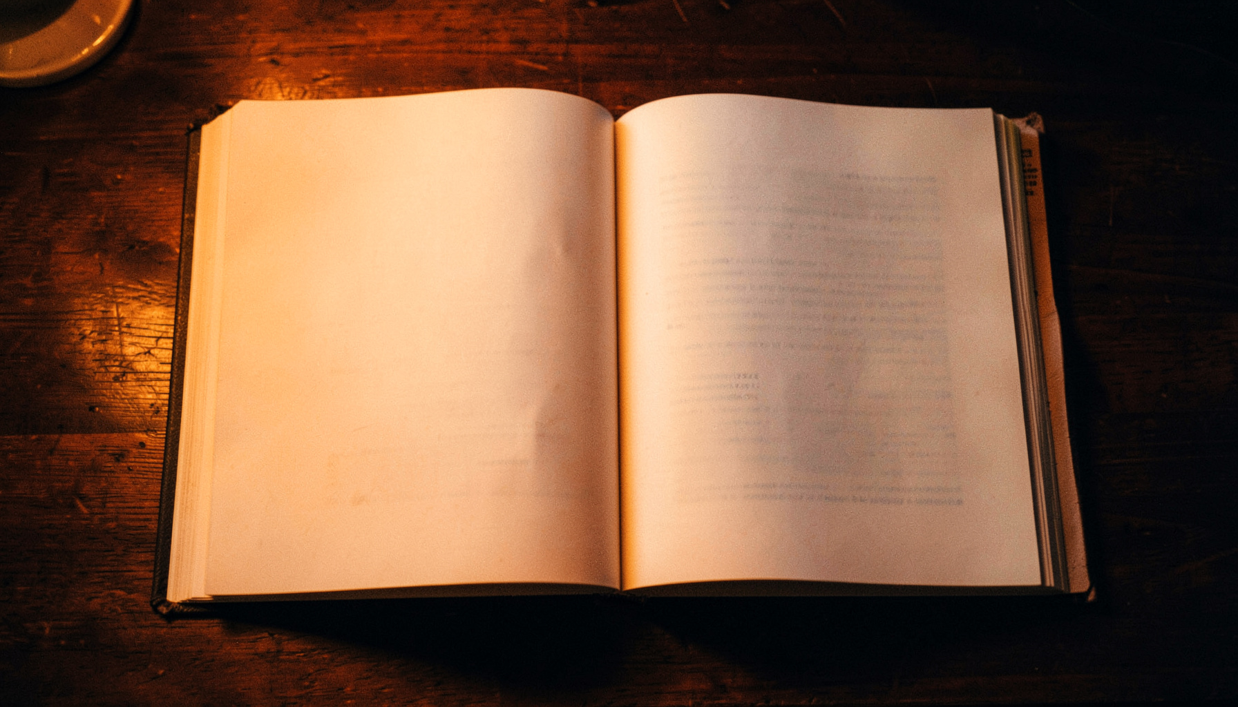 A warm-toned overhead shot of an open book on a dark wooden desk, soft lamplight pooling across the pages, deep browns and muted golds.